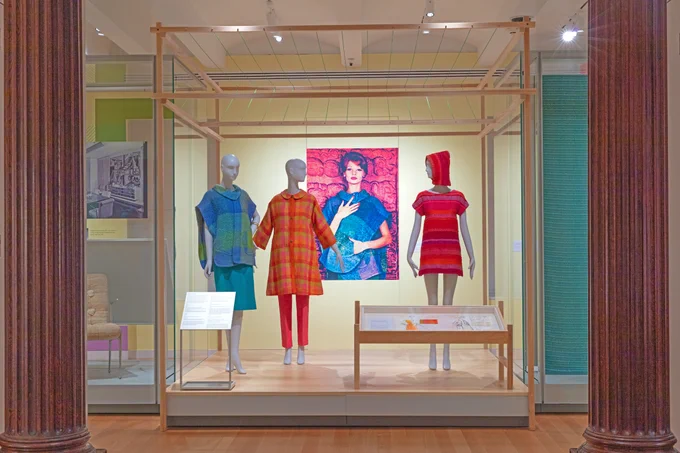 Three mannequins stand inside of a glass box wearing blue, green, orange, and pink garments designed by Dorothy Liebes. In between the mannequins is a vintage editorial photograph featuring one of the garments. 
