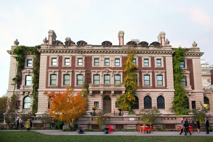 An exterior view of the Cooper Hewitt, Smithsonian Design Museum and garden. There are five rows of green and yellow ivy running up the building and small tree with orange leaves. There are red, black, and white tables and chairs, and groups of people in fall and winter clothing standing around the garden under a light blue sky.