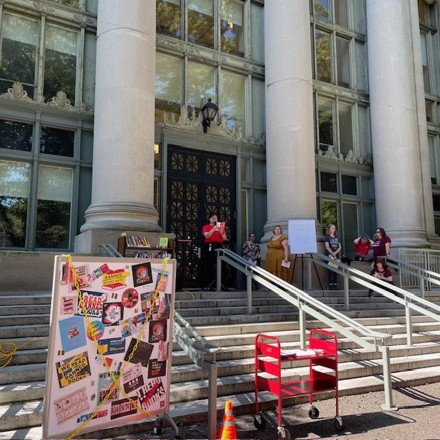  An HLS Library staff member stands at the top of the steps of Langdell Hall. She holds a microphone and reads aloud from a banned book. Several people stand to her left. In the foreground is a colorful sign advertising Banned Books Week. 