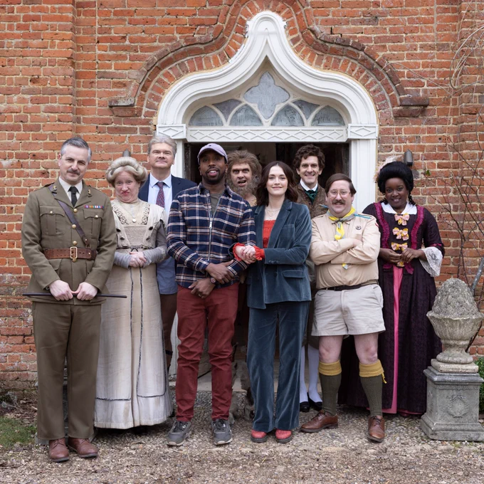 The cast of Ghosts assemble outside a door on the Button House set, smiling.