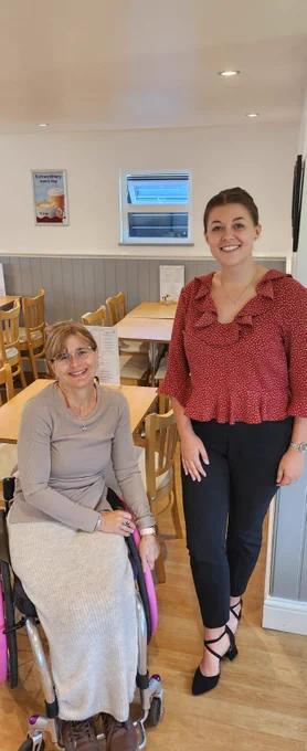 Councillor Amanda Worne and Ellen Ilott from AccessAble pictured next to each other in a café. Amanda is a wheelchair user and is wearing a light brown jumper with a long cream skirt and brown boots. She has blonde hair that is tied up and glasses. Ellen is standing next to Amanda and is wearing a red blouse with white polka dots, black trousers, and black heels. She has brown hair that is tied up.