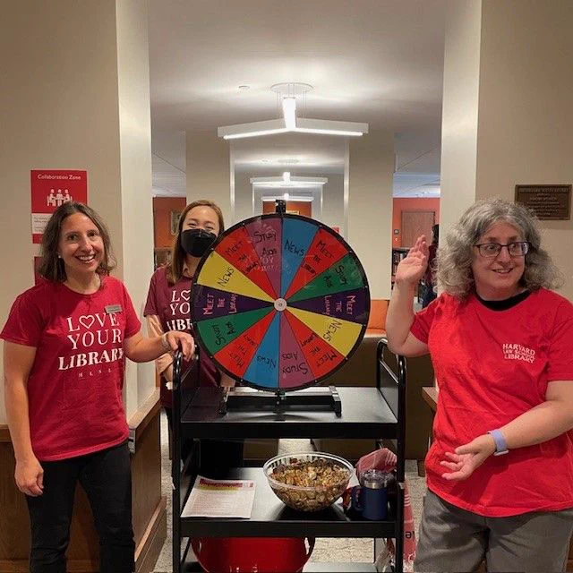  Three library staff members wearing "Love Your Library" t-shirts stand around a colorful wheel, which students could spin to learn about library services and research databases.