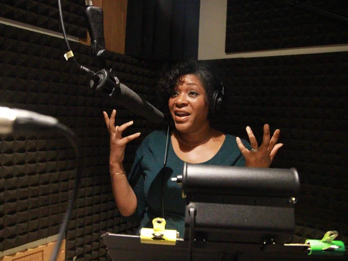 HUMAN POWERED co-host Dasha Kelly Hamilton wears headphones, and speaks into a microphone in a recording studio. Her hands are raised in a gesture for emphasis.