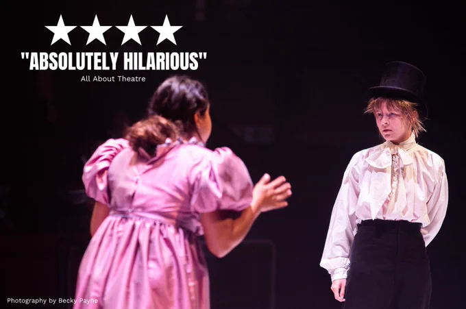 Anshula Bain and Heather Forster in We Could All Be Perfect. Photo by Becky Payne. A young woman dressed in a billowing shirt and black top hat stands and looks confused. In the foreground, another young woman wearing a pink regency dress is explaining something to them. 

Quote on top of image reads FOUR STARS "ABSOLUTELY HILARIOUS" (All About Theatre)