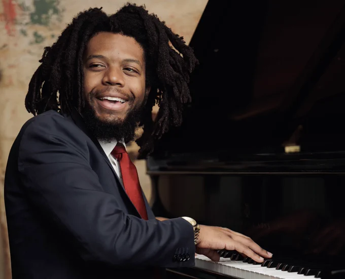 Sean Mason playing the piano while wearing a suit and tie. They are smiling. 