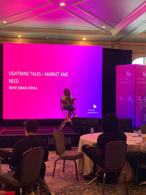 A Picture of Irene Mbari-Kirika seated on a chair on the Remarkable Stage. Behind her, there's a presentation written Lighting Talks - Market and Need, Irene Mbari-Kirika. There are people facing the stage and listening to her.
