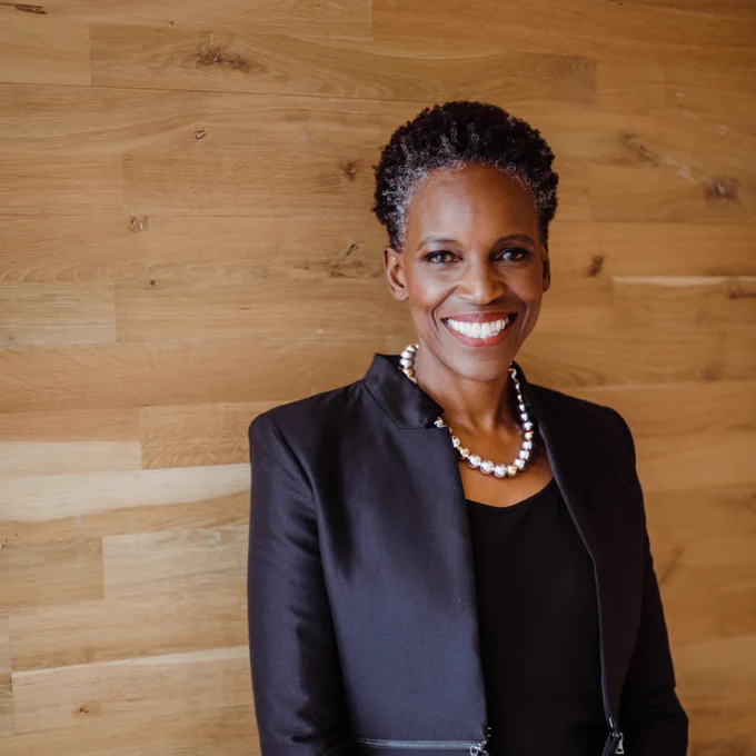 Photo: A black woman with short hair wearing a black blazer over a black shirt with a necklace, smiling for a portrait in front of a wooden shiplap wall.