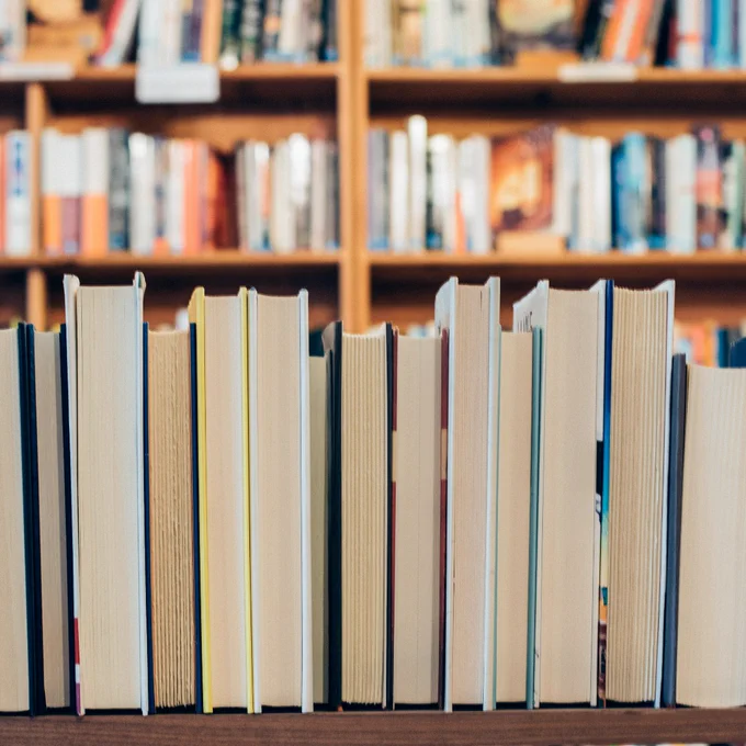 Photographs of a row of books with the pages showing. They are on a dark brown shelf, and in the background are brown bookshelves with books on the shelves. 
