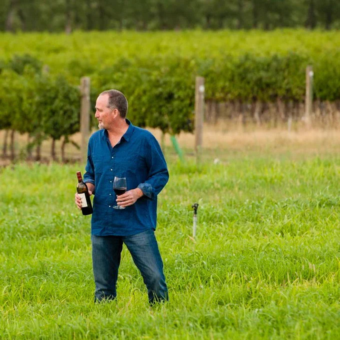 A person stands in a vineyard holding a glass of red wine in one hand and a bottle of wine in the other.