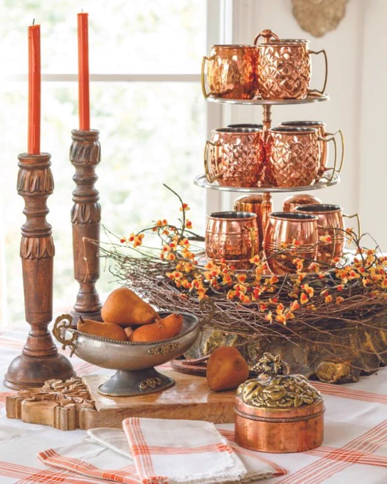 Second: A tabletop vignette consisting of orange tapers in copper candlesticks, copper mugs surrounded by branches, an orange-and-white plaid tablecloth with matching napkins, copper accessories, and a silver bowl holding pears.
