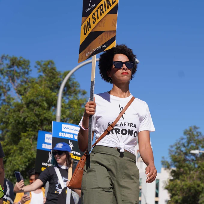 A person on the picket line leads the charge while wearing a SAG-AFTRA STRONG shirt and holding a SAG-AFTRA ON STRIKE sign. Many people are walking behind them.