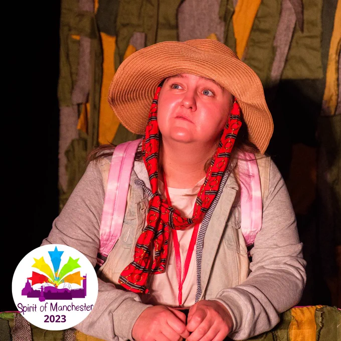 A performer is on stage playing Shadow Girl. They are delivering a monologue to an audience (out of shot). They look worried. There is a graphic image in white circle stamp which has the Spirit of Manchester 2023 logo in it. 