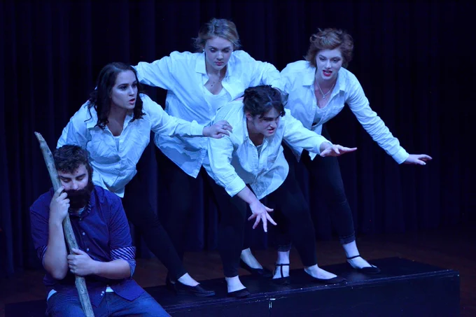 5 actors performing on a dimly lit stage. The person most leftward in the photo is wearing a blue top, while the others are wearing white tops.