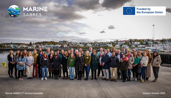 The Marine SABRES consortium stands in front of the harbour in Hafnarfjörður, Iceland opposite the Hafrannsóknastofnun (MFRI) building. The Marine SABRES logo is in the top left, and in the top right is the flag of the European Union with the text 'Funded by the European Union.' In the bottom right the text reads "Marine SABRES 2nd General Assembly, Iceland, October 2023.'