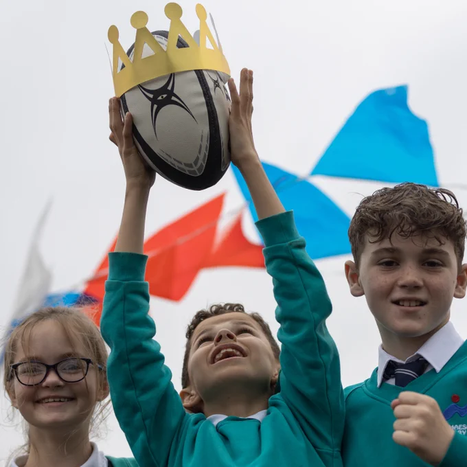 3 young Welsh pupils, one raising a rugby ball in celebration.