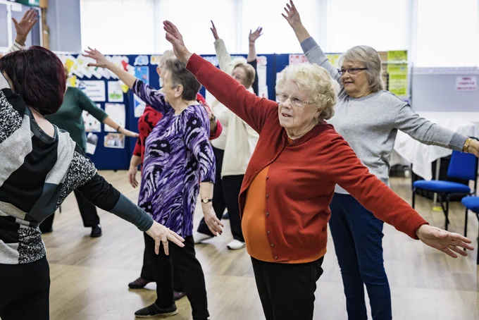 A large group of older adults are dancing in a community centre. Their arms are outstretched diagonally as though reaching to the sky and towards the ground. They are taking part in a Dance On session for older adults, run by darts in Doncaster.