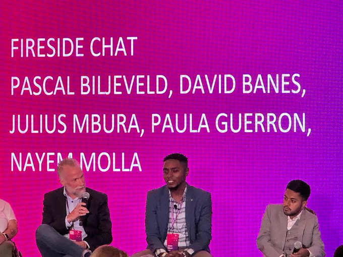 A picture shows Pascal Biljeveld, Julius Mbura & Nayem Molla seated near each other on a remarkable stage. They are all holding microphones. Pascal is speaking into his microphone and facing Julius Mbura and Nayem Molla on his left side.