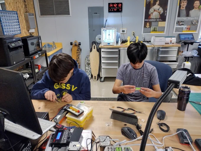 Zwei Kinder sitzen gebeugt an einem Schreibtisch in einem Maker-Space. Auf dem Tisch liegen Kabel und Werkzeug, im Hintergrund Computer, Lonboard, Drucker und weitere elektronische Geräte. 