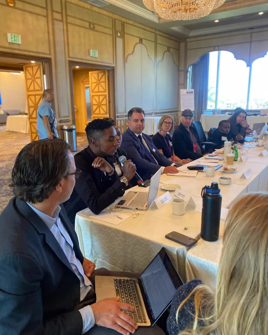 A picture of Julius Mbura holding a microphone and speaking during the AT Scale ideation workshop. He's seated at a table with other people, and they are facing him and listening to him.