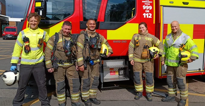 Five firefighters in fire kit stand beside a fire engine with the fire station name 'Ward End' on the door