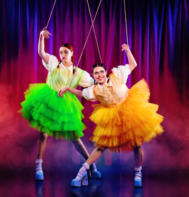 Maddy and Marina Bye, who make up Siblings. They are two women, wearing plaits, circus style make up, puffy chiffon orange and green skirts and chunky high heels. They have their arms in the air held by strings like puppets and make exaggerated clown facial expressions. Behind them is a purple curtain, as if they are on stage.