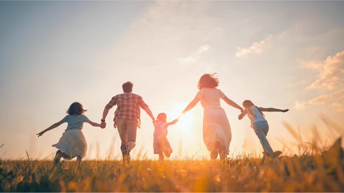 Image of a family made up of two adults and three children holding hands and starting to run in an open field towards a sunrise.