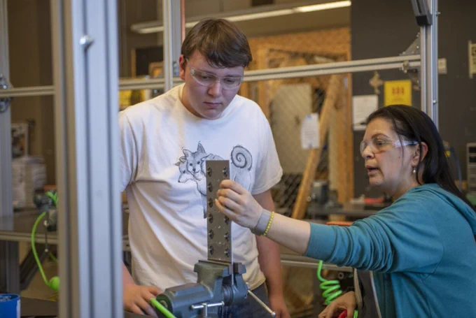 A teacher explaining a project using a piece of metal to a student inside of a shop class. 