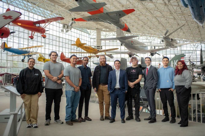 A group photo of 10 people standing inside of a museum with airplanes. 