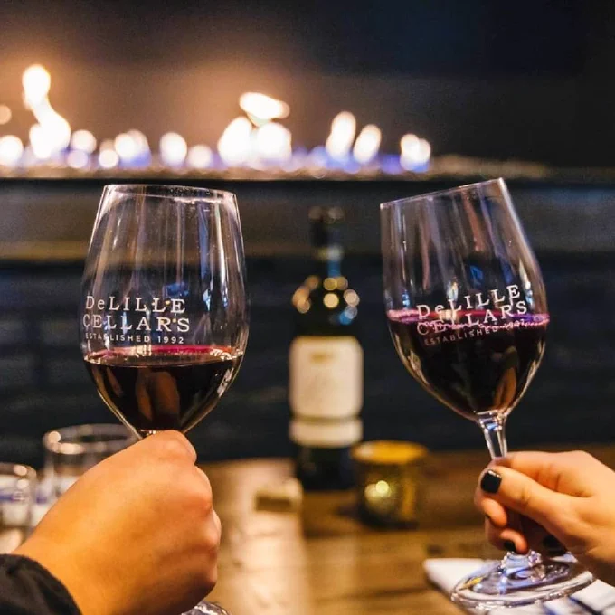 Two hands hold wine glasses with red wine in front of a fireplace.