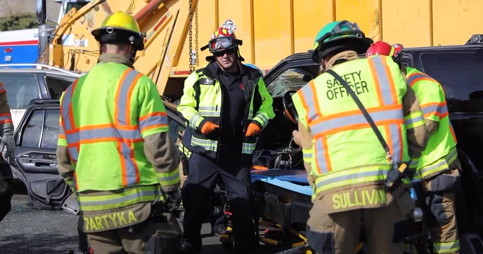 Three first responders in high-visibility vests and safety gear working together in front of a staged crash.