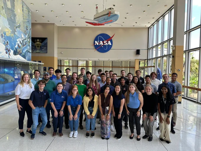 A group of about 35 interns stand in a hall at Johnson Space Center posing for a group photo. The NASA insignia is on the wall behind the interns. The wall at left has a painting of a space shuttle and astronaut and a glass memorabilia case below. Credit: NASA