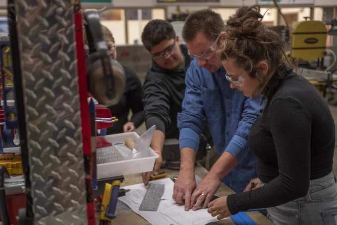 An image of two students getting instruction from a teacher inside of a shop classroom. The students and teacher are looking at a blueprint. 
