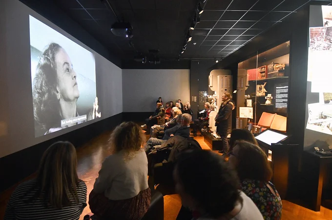 A group of people watch a video on a large film screen in a gallery.