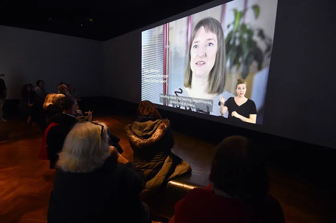 A group of people watch a video on a large film screen in a gallery.