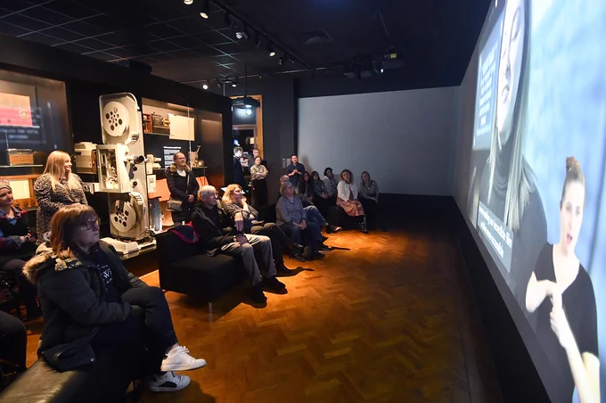 A group of people watch a video on a large film screen in a gallery.