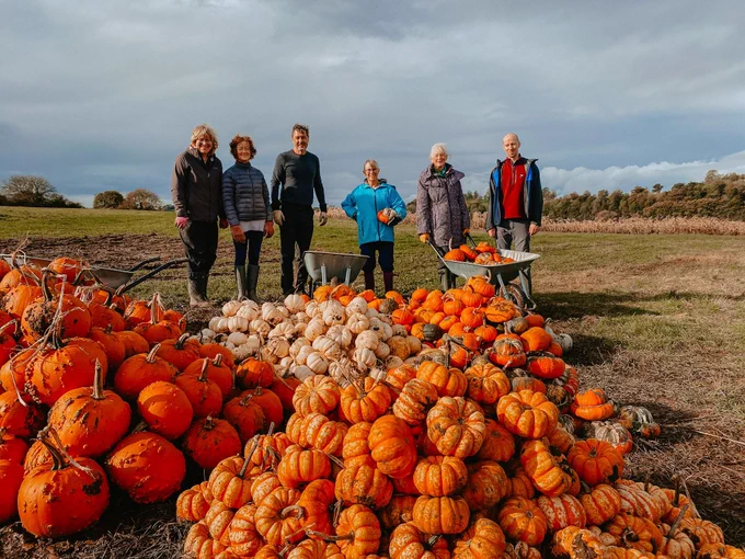 Six people pose next to a huge pile of gleaned pumpkins. They are smiling and the sun is out.