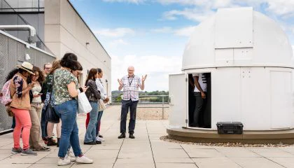 A crowd of students stood listening to a professor next to a rooftop observatory.