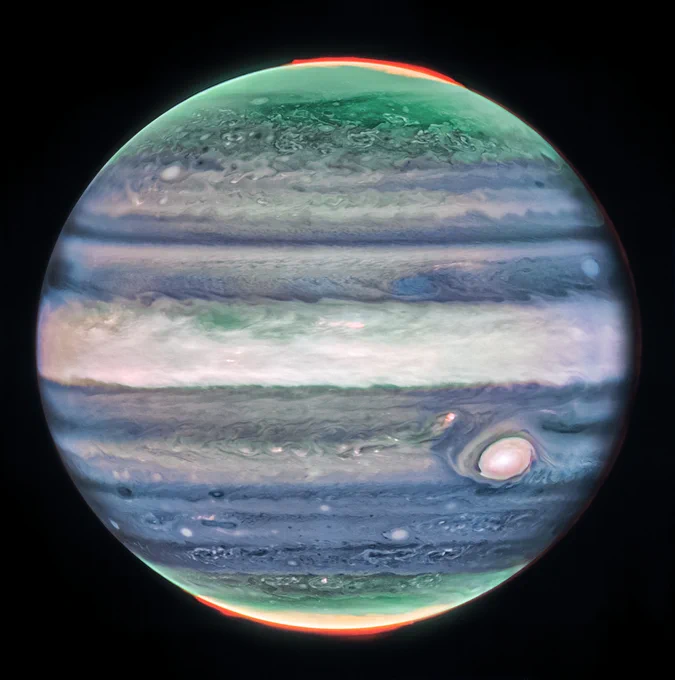 Jupiter dominates the black background of space. The image is a composite, and shows Jupiter in enhanced color, featuring the planet’s turbulent Great Red Spot, which appears white here. The planet is striated with swirling horizontal stripes of neon turquoise, periwinkle, light pink, and cream. The stripes interact and mix at their edges like cream in coffee. Along both of the poles, the planet glows in turquoise. Bright orange auroras glow just above the planet’s surface at both poles.