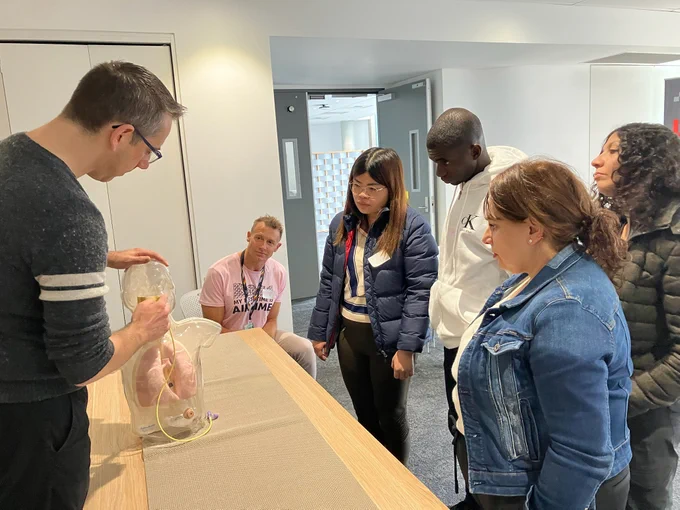The photo shows a group of people taking part in the pilot training session of SUCCEED's Nasogastric Feeding Tube course, using Adam,Rouilly's AR331 Hungry Manikin Paediatric Feeding Trainer.