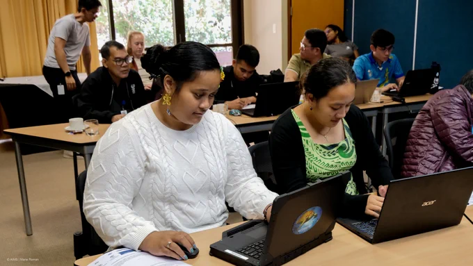 Two Samoan women are on laptops with others behind in a classroom setting.