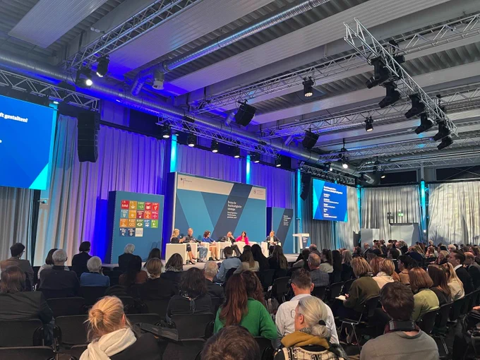 Es ist ein großer Raum mit Menschen im Publikum zu sehen. Diese hören einer Podiumsdiskussion zu. Im Hintergrund sind die 17 SDG-Icons zu sehen und mit verschiedenen Blautönen designte Aufsteller und Folien, die deutlich machen dass das Bild auf der Auftaktveranstaltung zur Weiterentwicklung der deutschen Nachhaltigkeitsstrategie aufgenommen ist.