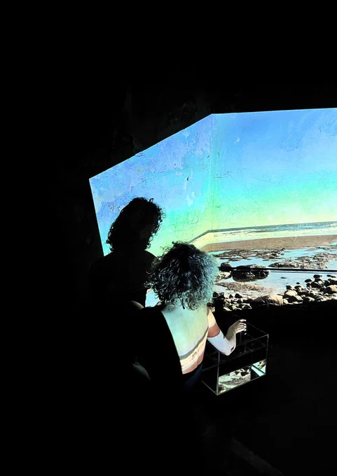 A white woman with short curly hair sits in a dark room facing away from the camera. A projection is being played onto her of a beach and the sea. She reached her arm out to touch a fish tank to her right.