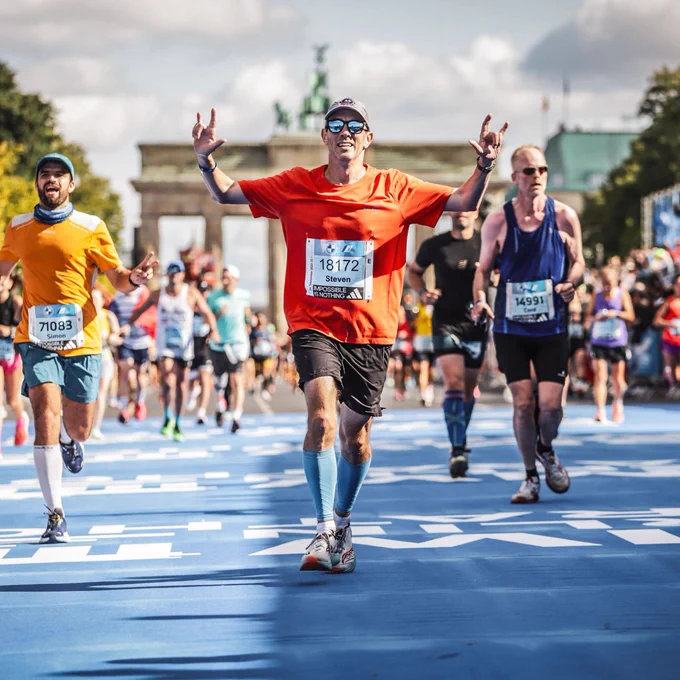 A man in the center of many runners signs, "I love you," with both hands while running to the finish line of the 2023 Berlin Marathon. In the background is the Brandenburg Gate.