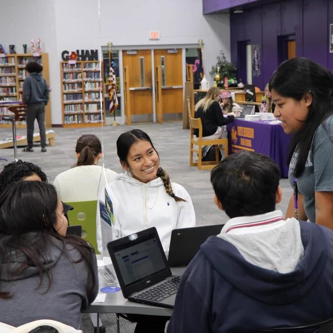 Four students sitting at a table in the media center talking to an adult.