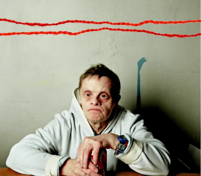 Judith Scott sits at a desk with her hand resting over a soda can. Two pieces of decorative thread run across the top of the photo

Image Source: Art21.org