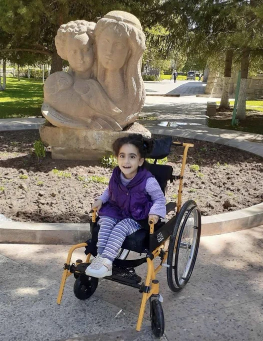 A young girl dressed all in purple with curly dark brown hair is sitting and smiling in a yellow wheelchair. She's outside by a statue of a woman and a dove.