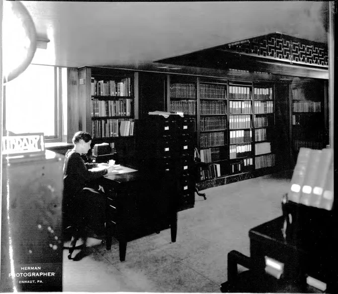 A person sits at a desk writing surrounded by bookcases and filing cabinets. 