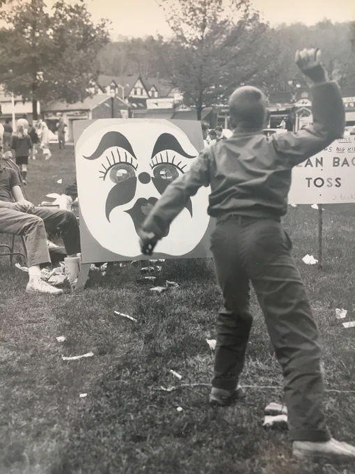 A young boy is throwing a ball at a carnival. The boy is aiming for the mouth of a clown painted on a wooden board. 