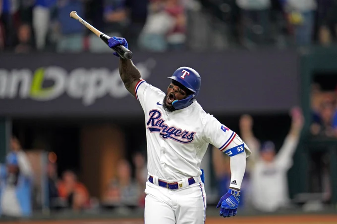 Adolis García raises his bat above his head with his right hand as he slams it to the ground after hitting a home run during Game 5 of the ALCS on Friday. He is wearing a white Texas Rangers home uniform with blue and red lettering.