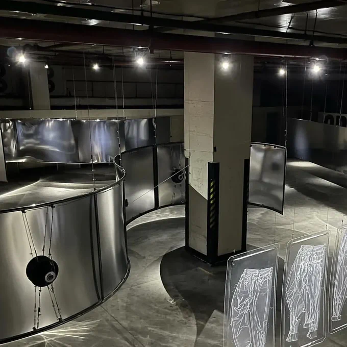 A photo of an art installation in an underground car park space. There is a row of spotlights at the top, a large strip of silver curves around a white pillar. It has round speakers strapped to the back of it. Another curved sheet sits opposite. In the foreground are the impressions of trousers, cast in vacuum formed, clear plastic.  

Image: No Tracksuits, No Trainers, 2023. © Hannah Perry.